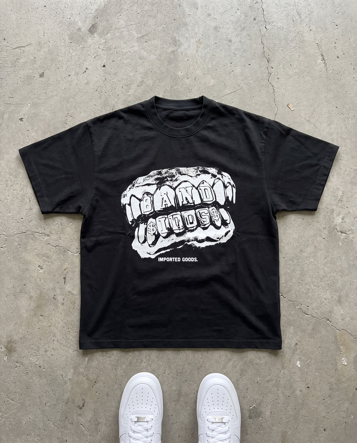BANDIDOS GRILL TEE laid flat on concrete – black tee with white BANDIDOS grillz graphic and IMPORTED GOODS text