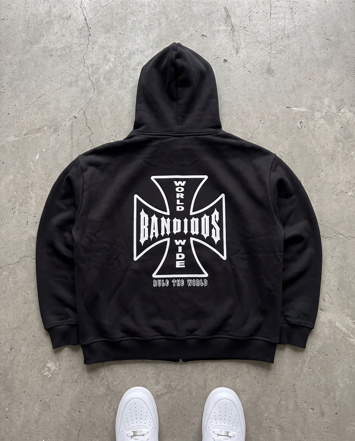 BANDIDOS STAPLE HOODIE back laid flat on concrete – black zip hoodie with white BANDIDOS cross graphic